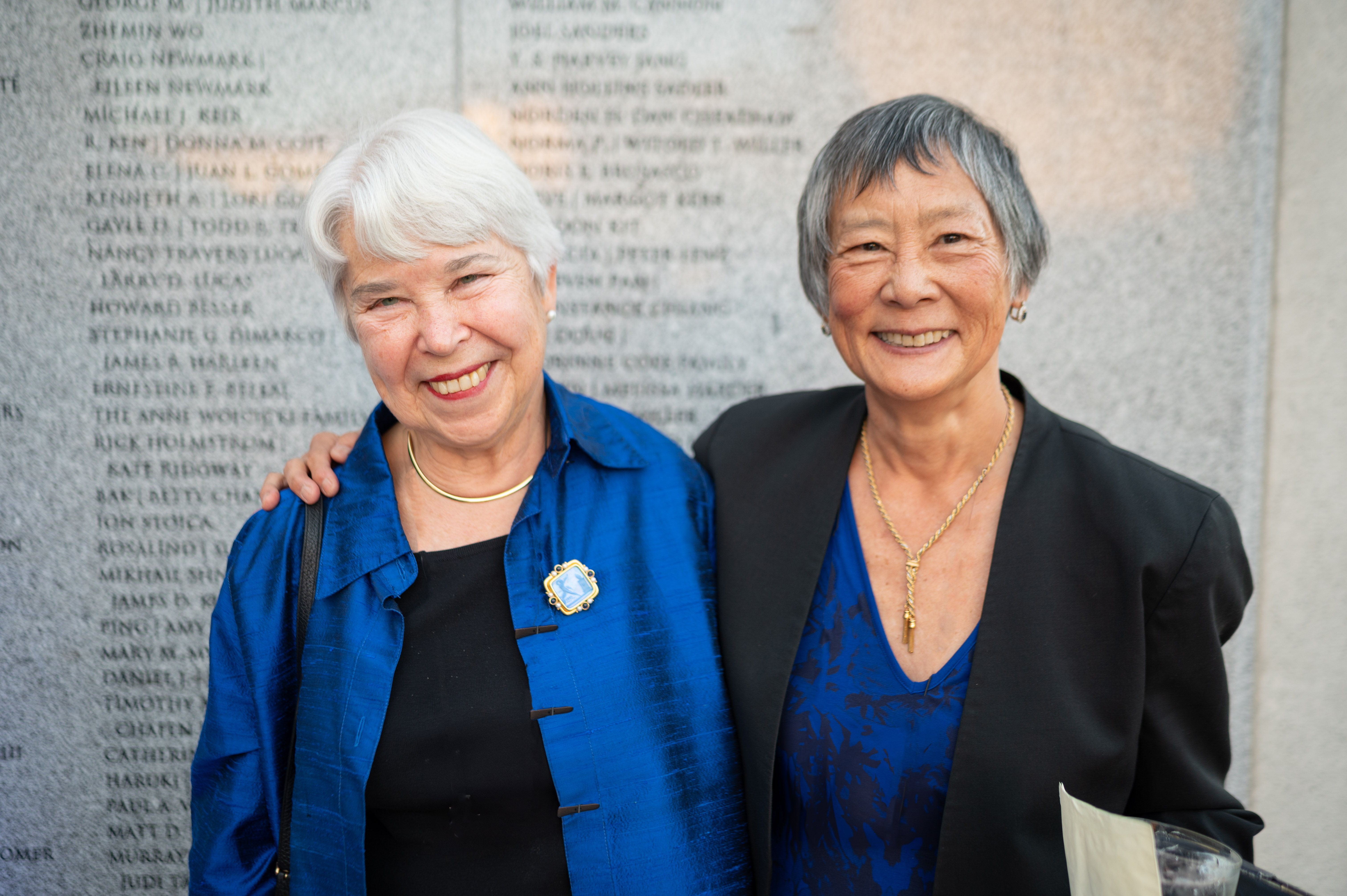 Photo of Chancellor Christ in a black top and blue blazer and Nadine Tang in a blue top and black blazer standing and smiling in front a wall etched with donor names.