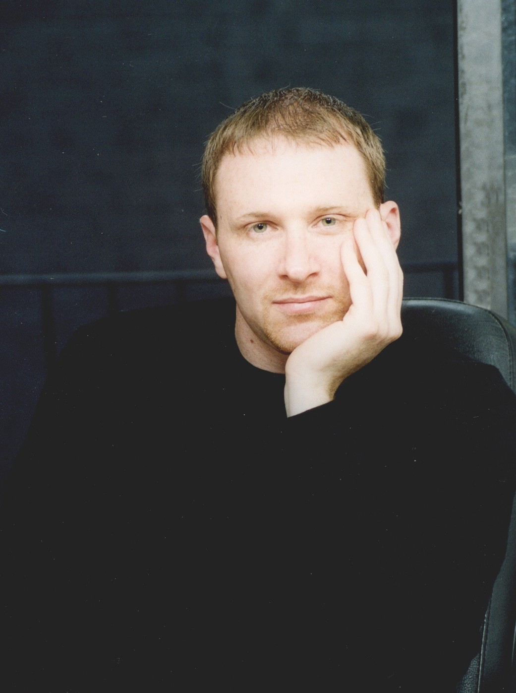 A photo of Christopher, who is wearing a black shirt and has his left hand cradling his face.