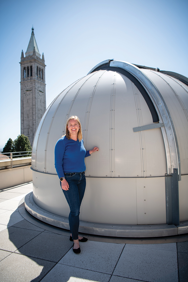 Photo of Courtney wearing a cobalt blue sweater and blue jeans, leaning against a round observatory with the Campanile in the distant background.