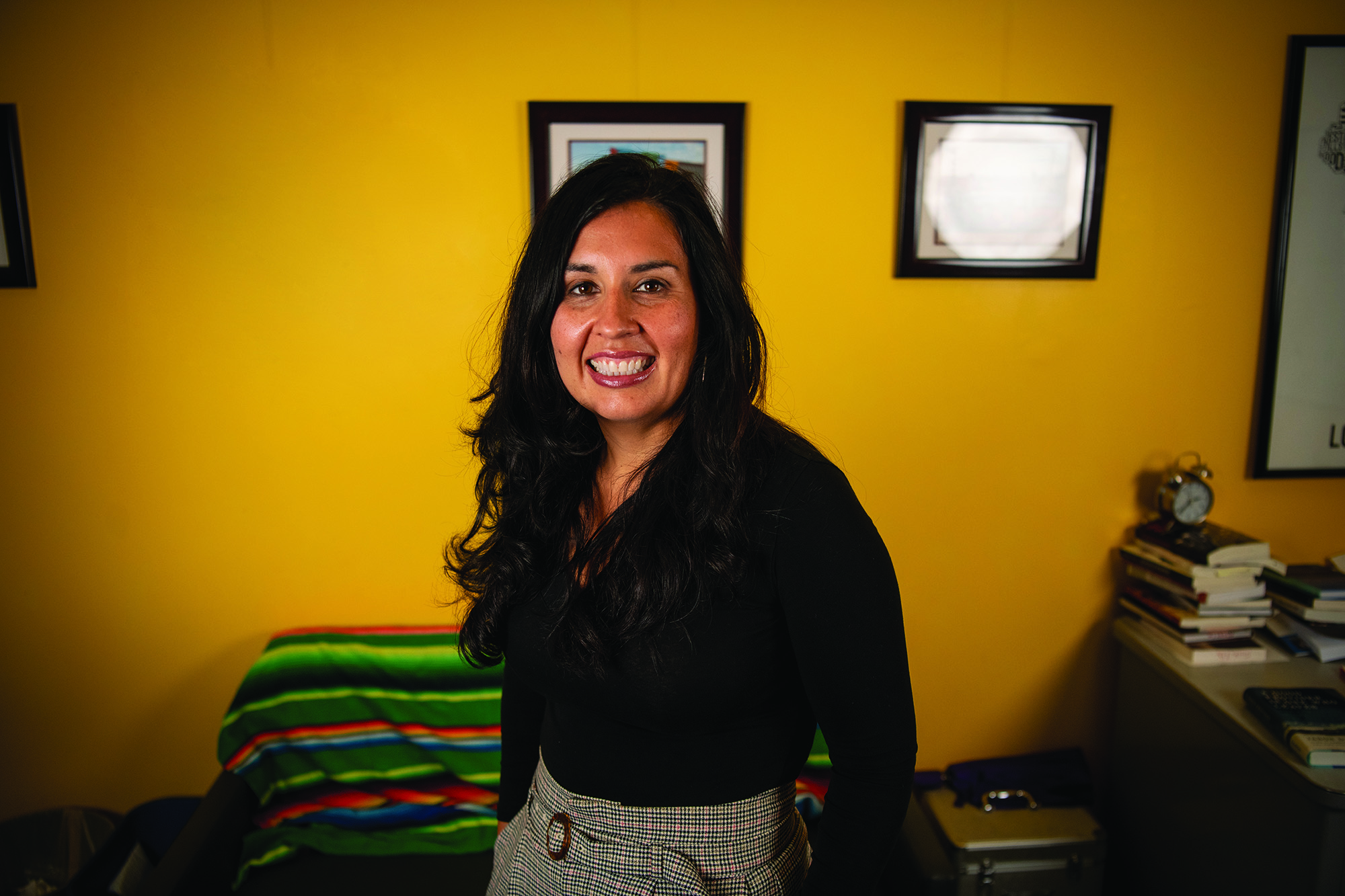 Photo of Cristina wearing a black top and neutral-toned plaid pants, smiling in front of a bright yellow wall.