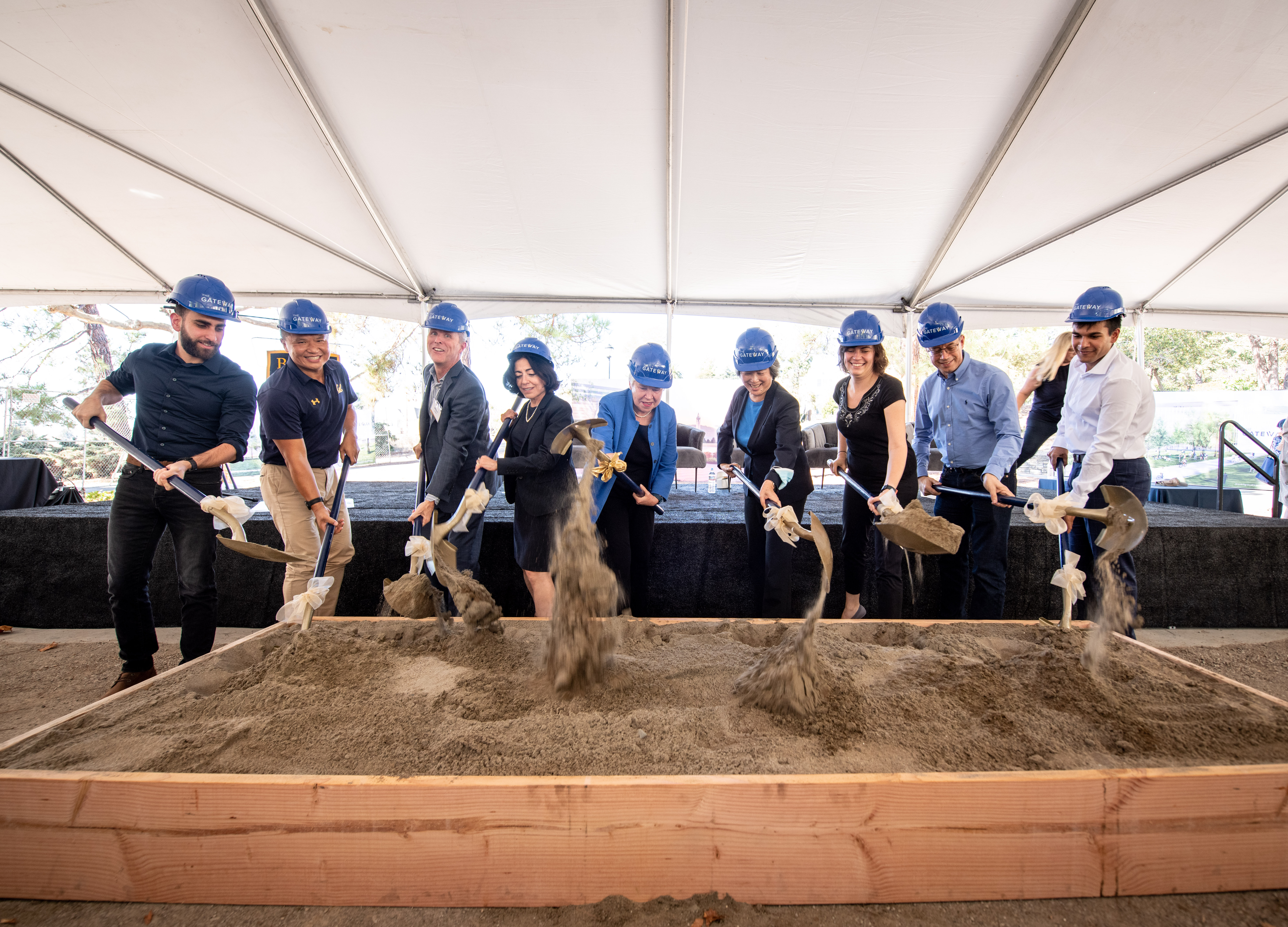 Photo of dig participants in blue hardhats digging into a sandpit and lightly tossing the sand.