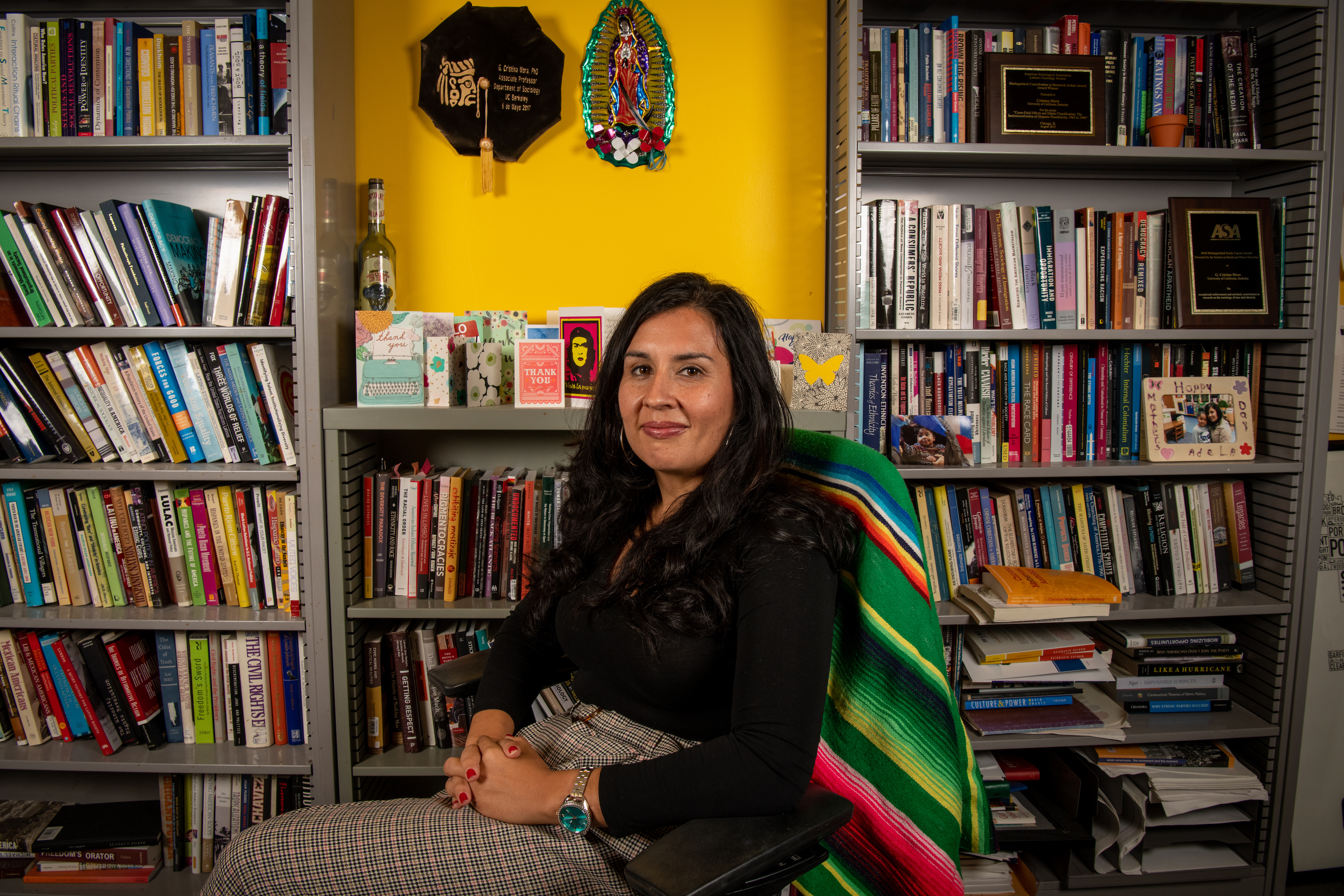 Photo of Professor Mora sitting in a brightly colored office surrounded by books, a Mexican blanket ont he back of her chair.