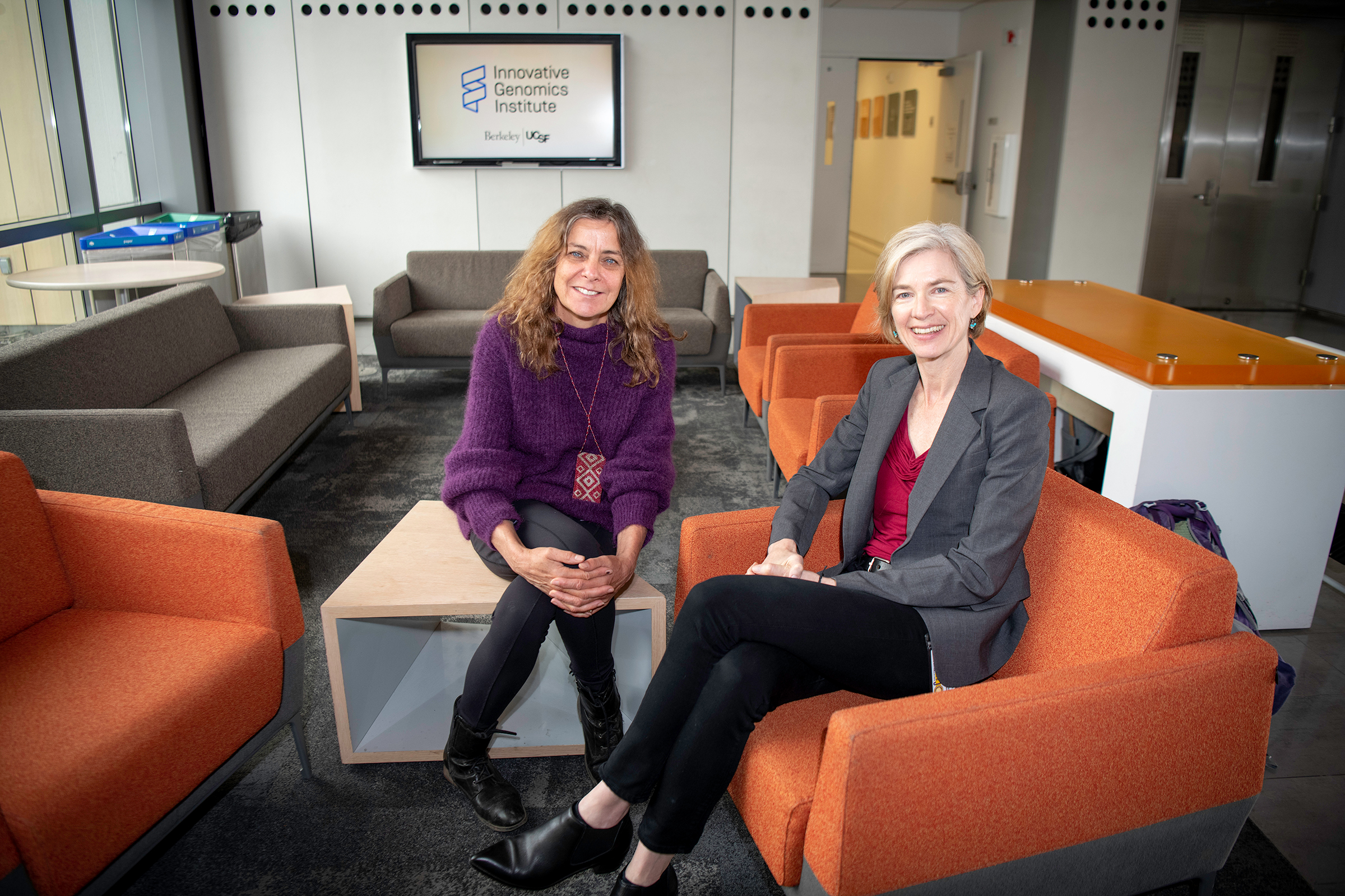 Jill is wearing a purple sweater and black pants, and jennifer is wearing a red top, gray jacket, and black pants. They are sitting in a lounge area with orange couches.