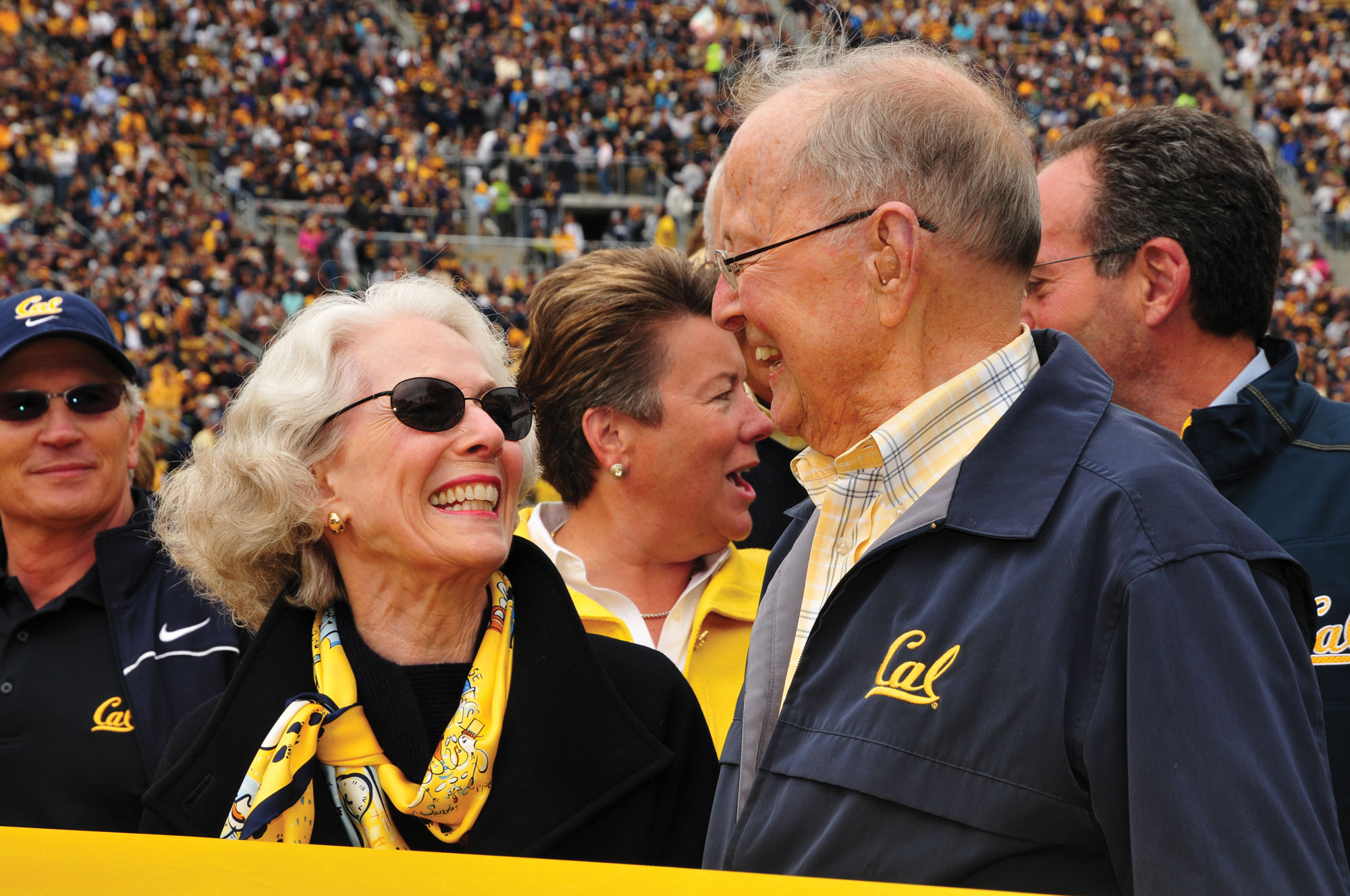 Photo of Sharon and Barc Simpson decked out in blue and gold, smiling at each other with full stands of Memorial Stadium in the background