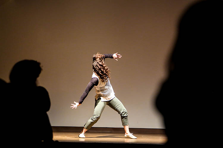 Photo of a dancer wearing street clothes standing with her feet spread wide and twisting back toward her left.