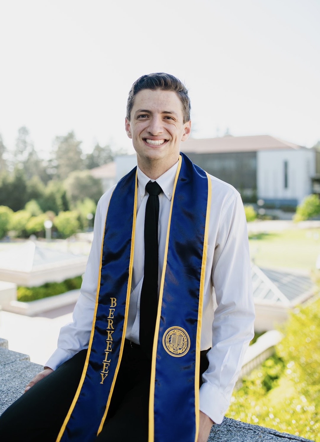 Daniel is smiling and wearing a white shirt, black tie, and a graduation stole. The East Asian Library is in the distant background.