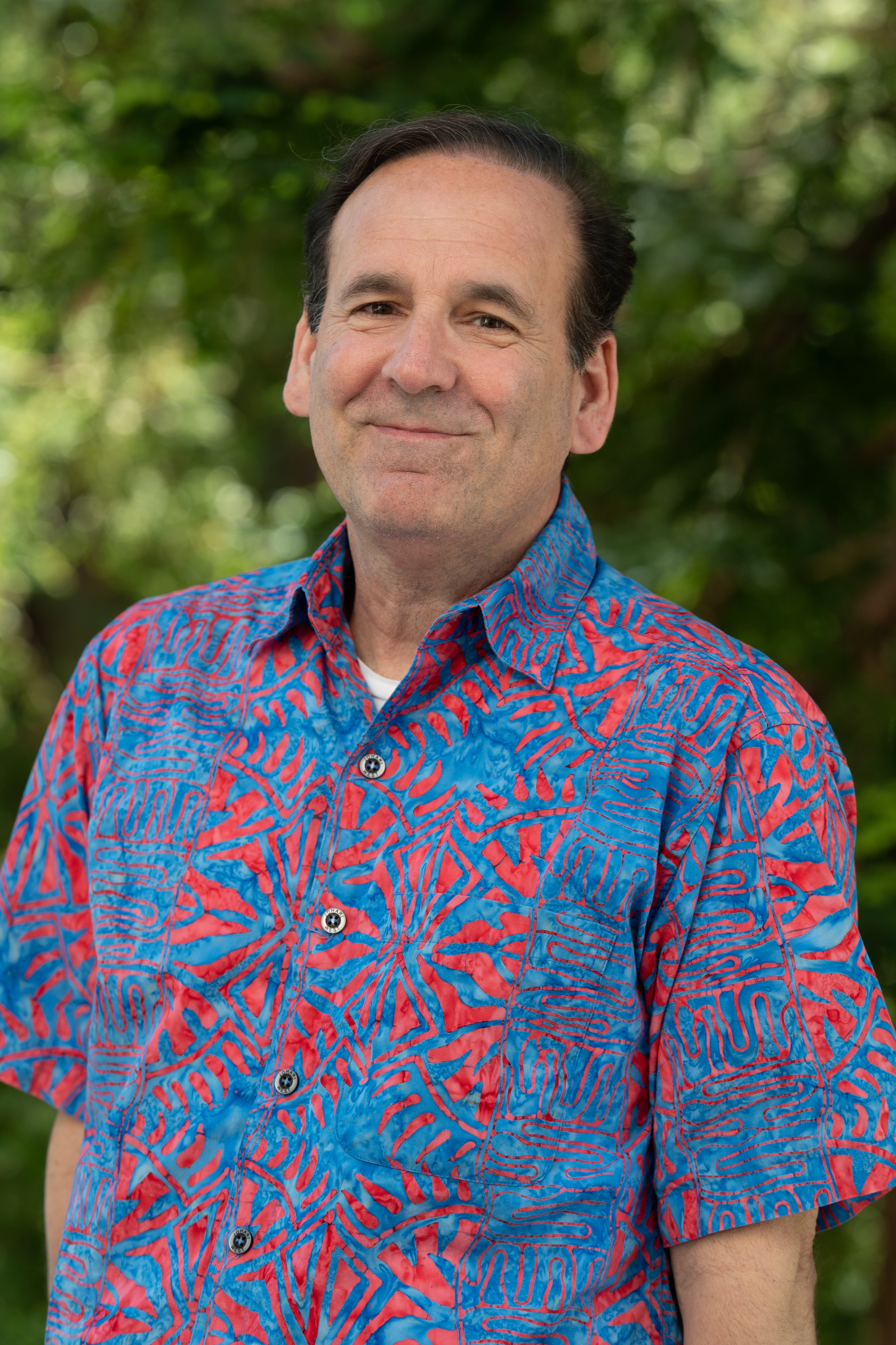 Photo of Darren in a blue and red batik-style print shirt standing with trees in the background.