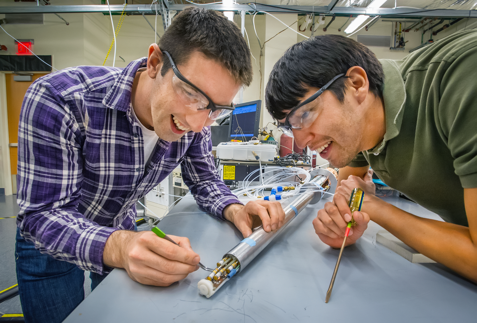 Photo of two male students, one in plaid and one in a green, short-sleeved shirt, wearing protective glasses and working with an engineering project.