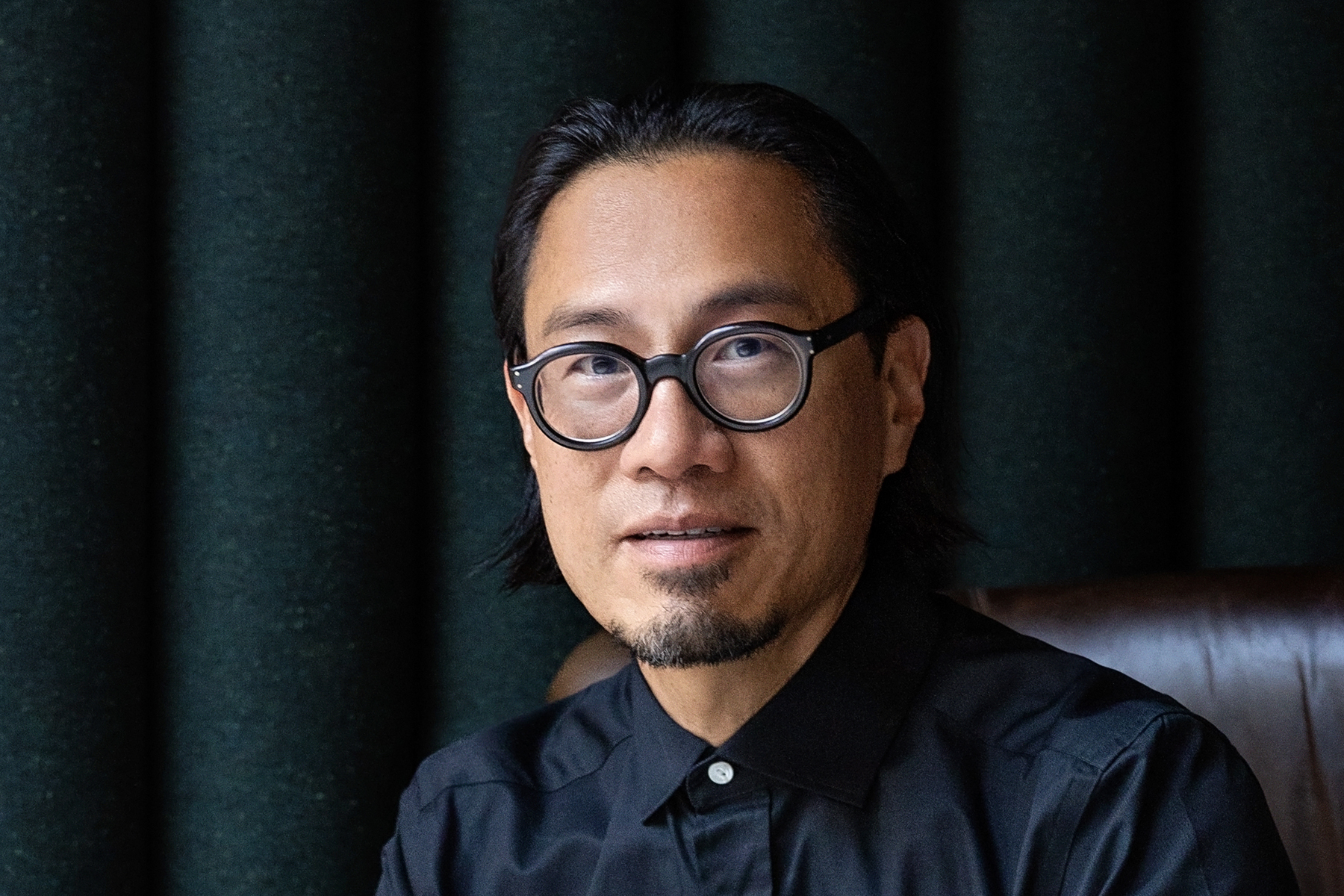 Headshot of Hao Ko '94, an Asian American man wearing a black top and round black glasses against a dark background