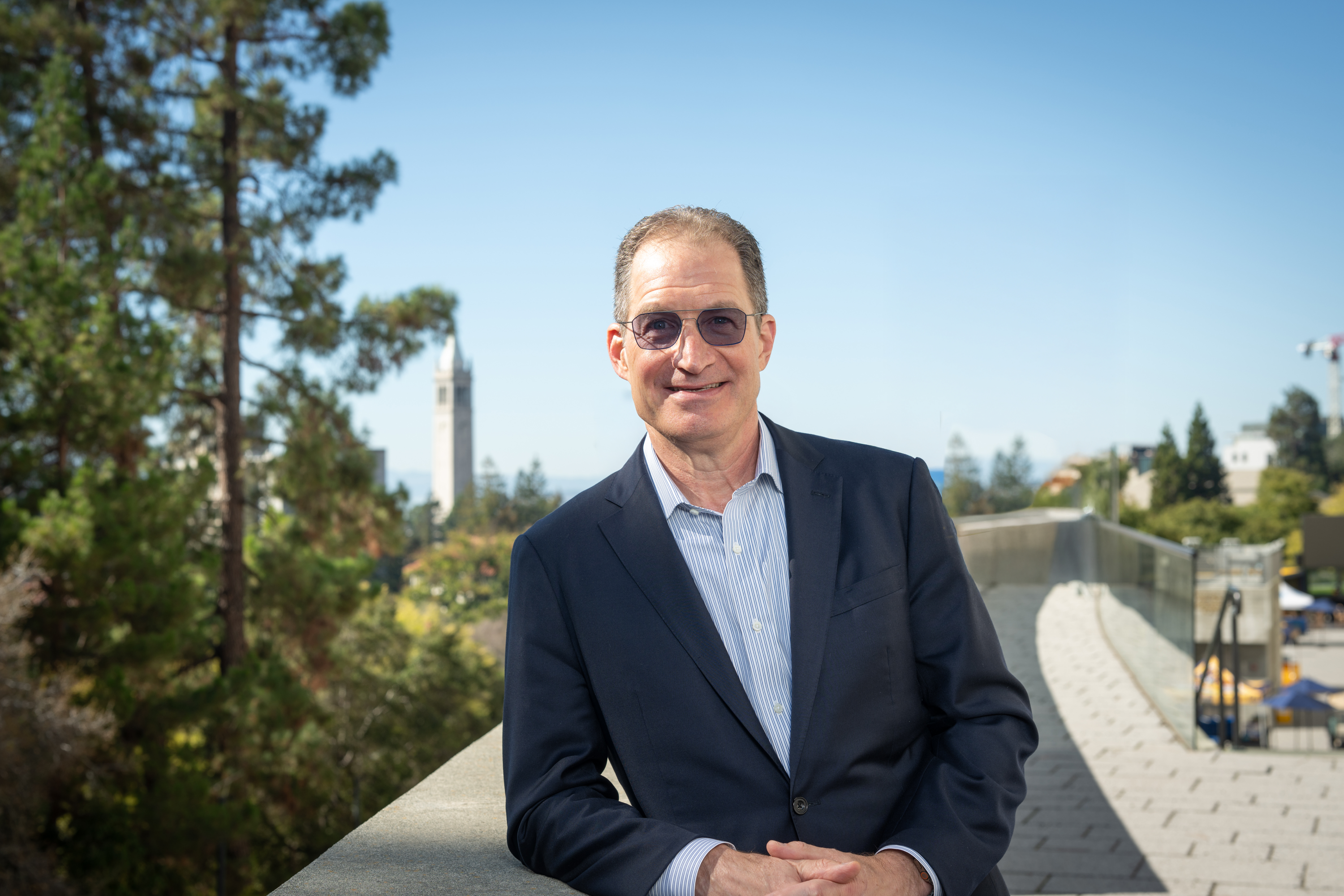 Photo of Eric in a dark suit leaning against a wall with the Campanile under a blue sky in the distance.