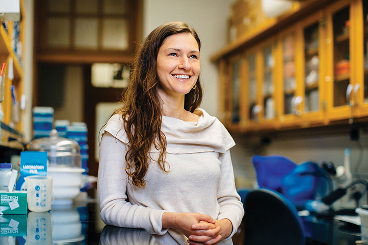 Photo of Erica with long brown hair and cream-colored top with a cowl neck, leaning onto a lab countertop.