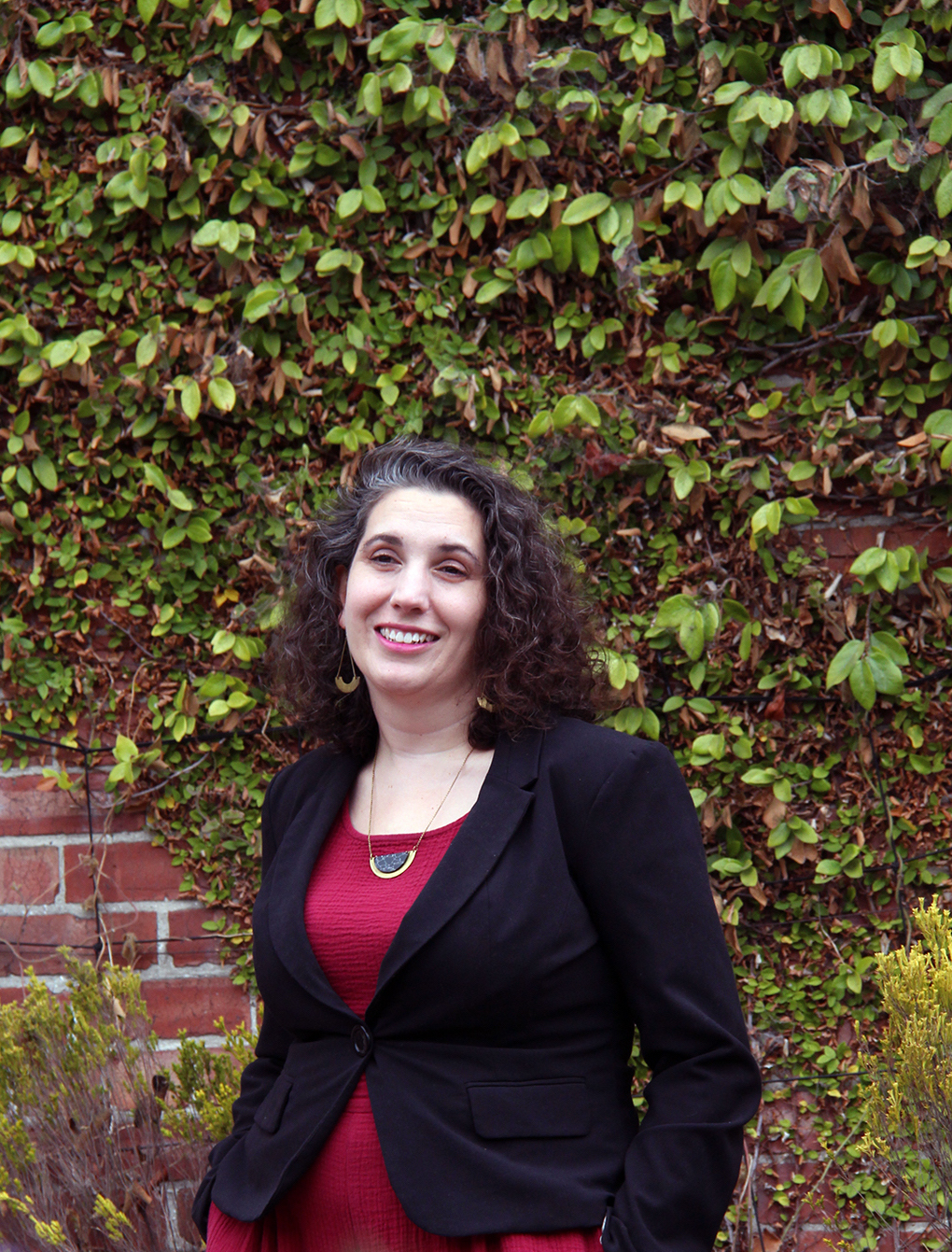 Photo of Evelyn standing against an ivy-covered brick wall, wearing a red top and black jacket. She is smiling and has long, dark, wavy hair.