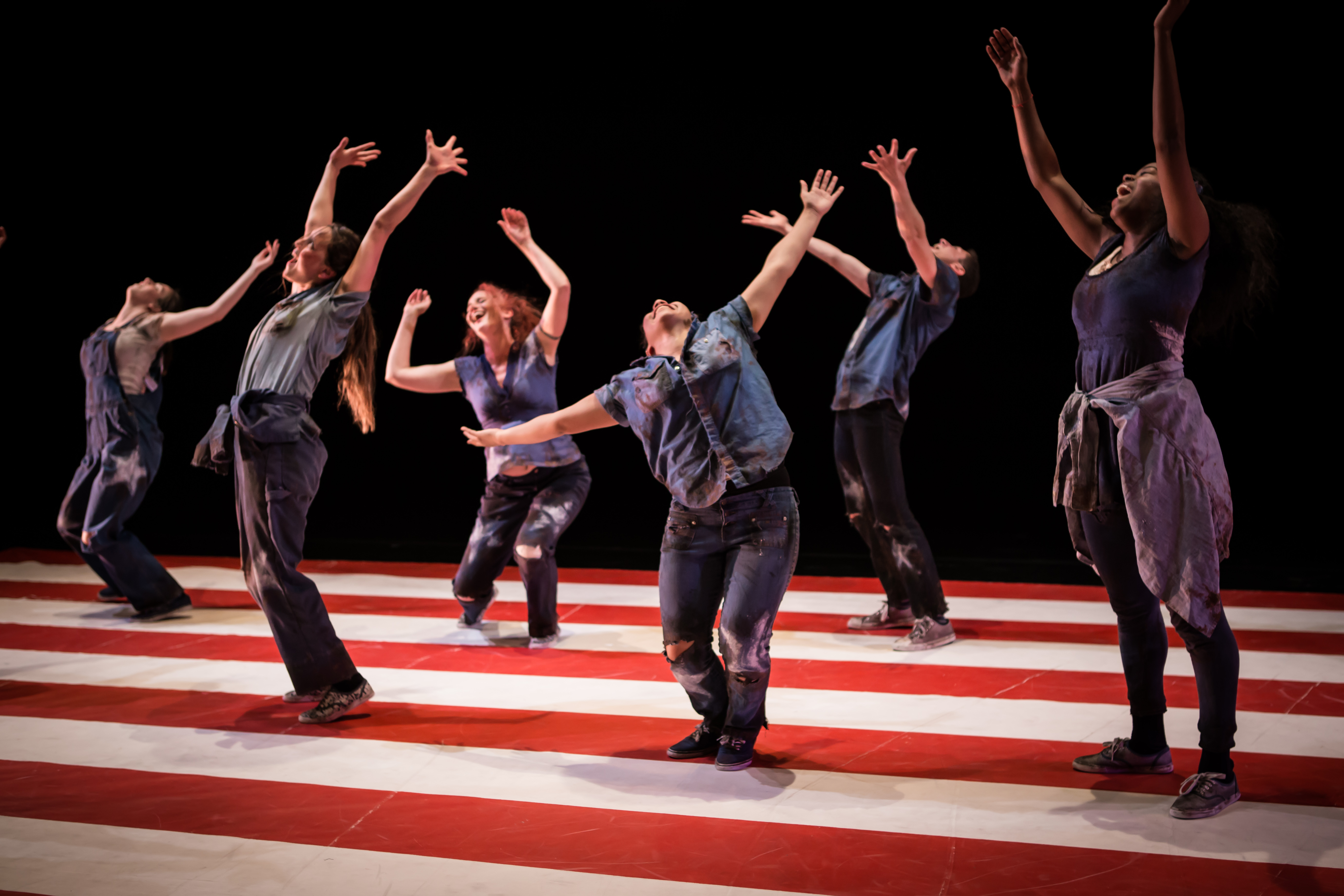 Photo of six dancers clad in blue work clothes, dancing on the red stripes of an American flag