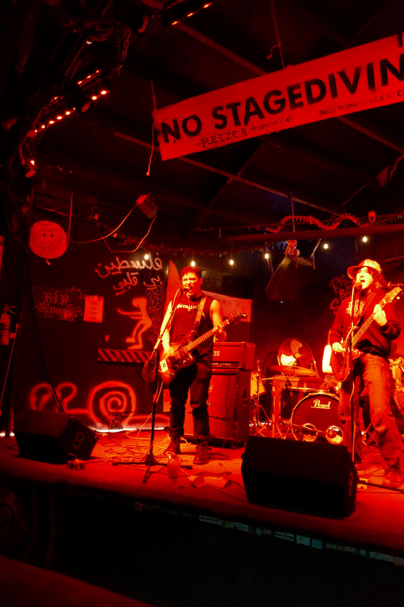 Photo of two singers/guitarists on a stage filled with music equipment under red lights.