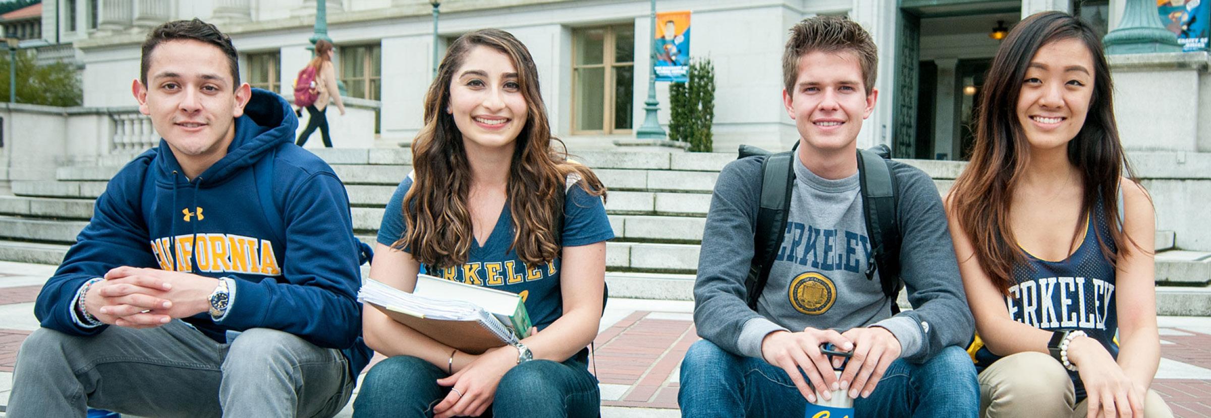 Photo of four students sitting on steps, smiling at the camera, all wearing UC Berkeley shirts of different kinds