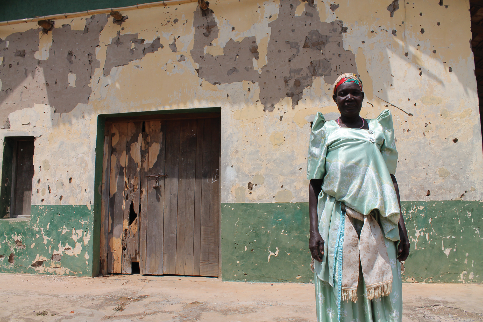 Photo of a woman in a green and white dress and colorful head wrap, staring straight at the camera unsmiling, standing in front of a weathered building.