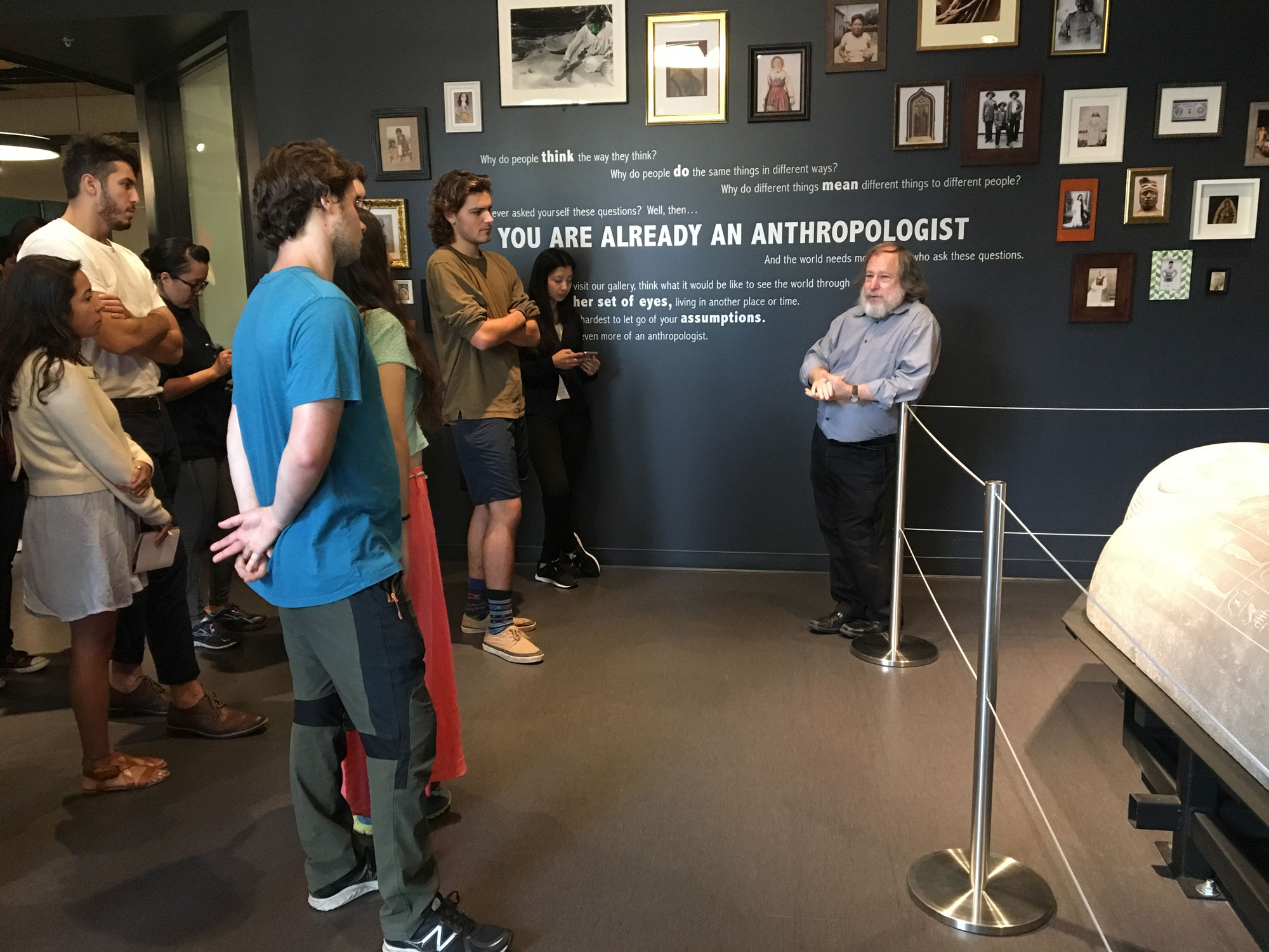 Photo of a Hearst Museum staff member and several students in front of a wall exploring the role of an anthropologist.