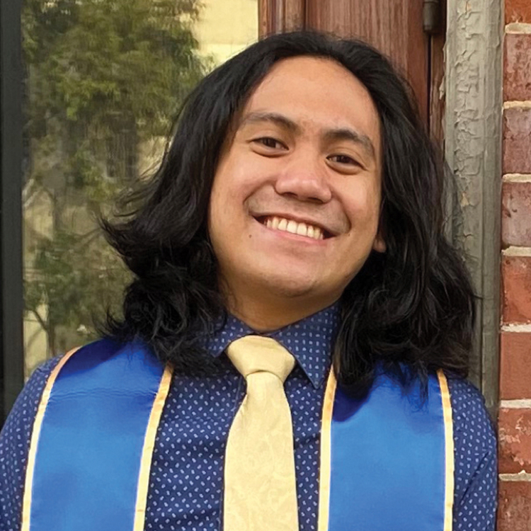 Photo of Ian with long, dark hair, a blue shirt and yellow tie, and his graduation stole.