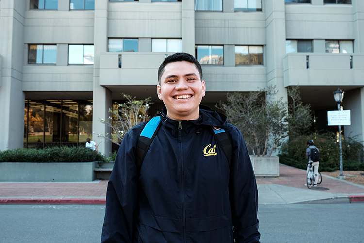 Photo of Ivan standing in front of a campus building, smiling and wearing a navy blue Cal jacket and backpack.