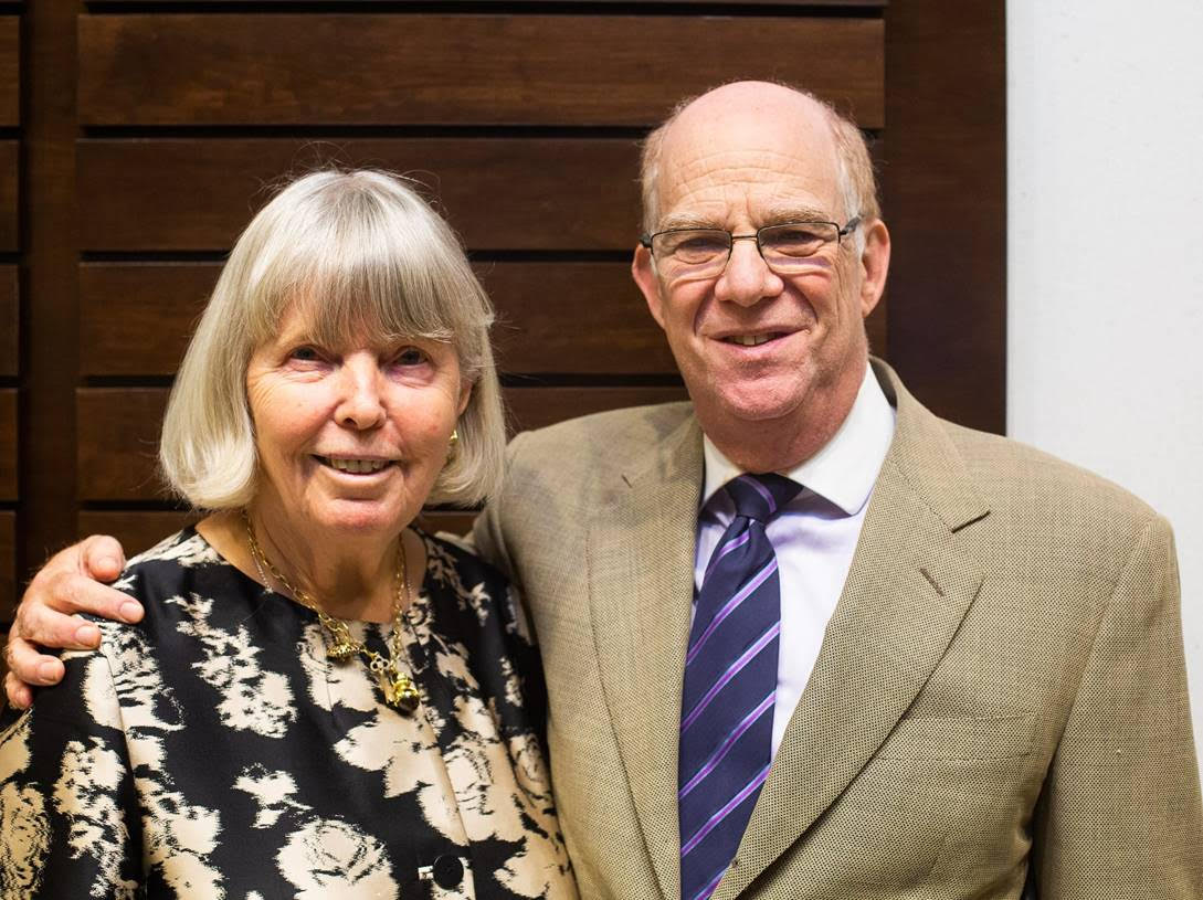 Photo of Kathleen, wearing a black top with white flowers, and James, wearing a blue and purple striped tie and pale olive green  jacket. He has his arm around her.
