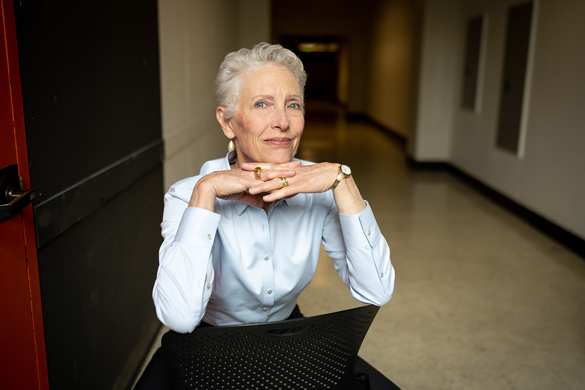 Photo of Jennifer sitting facing the back of a chair with her hands crossed beneath her chin. She has short gray hair and is wearing a white button-up shirt.