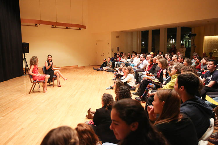 Photo of two women sitting in chairs on a stage and talking with a full, engaged audience.