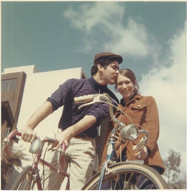 Jim, who's wearing a newsboy cap and striped shirt, leans in to kiss his new bride Gail, who's smiling at the camera. They're both on bikes.