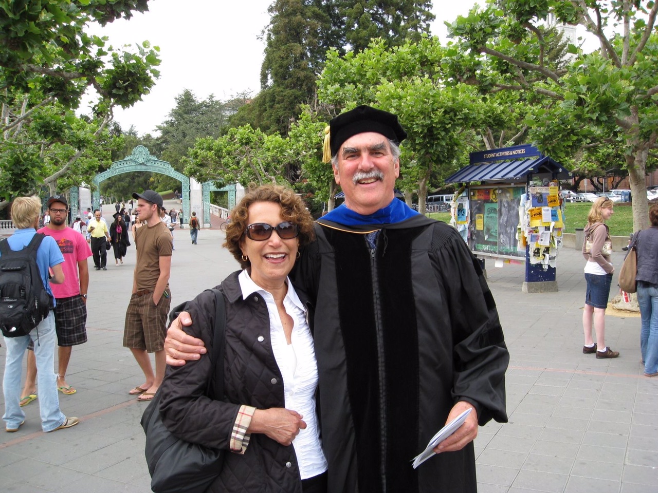 Jim, who is wearing his graduation cap and gown, hugs Gail. Sather Gate is in the background.