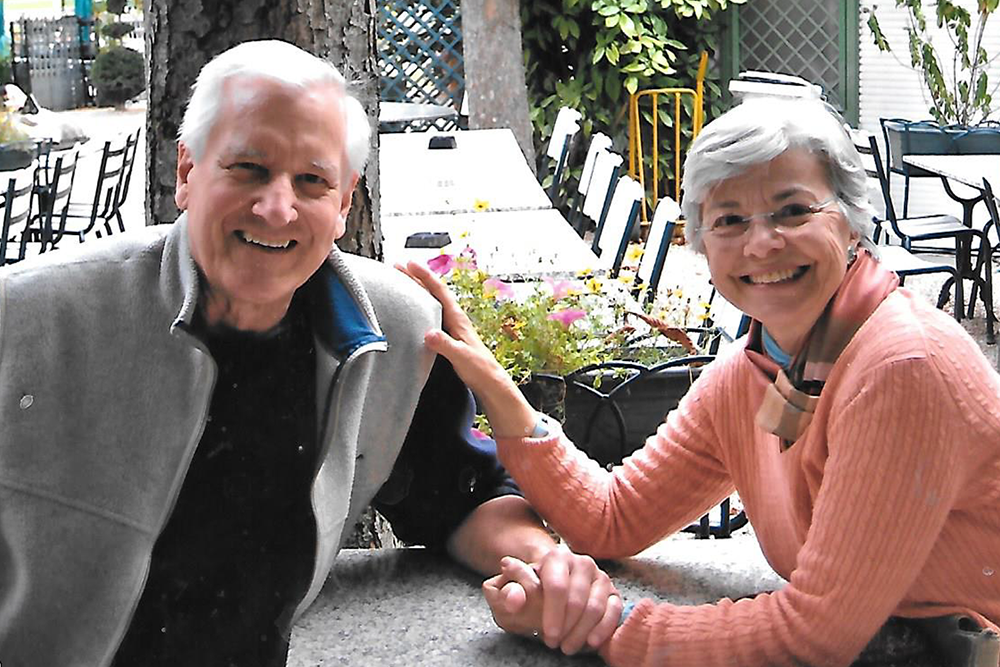 Photo of John (gray vest) and Beclee (peach sweater) sitting across the table from one another holding hands and smiling at the camera.