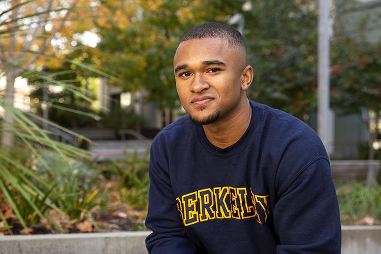 Photo of Kai wearing a navy blue Berkeley sweatshirt, sitting and smiling.