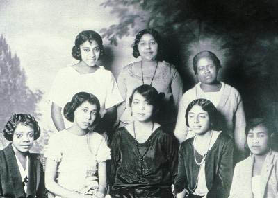 A historic black and white photo of eight Black women against a painted backdrop.