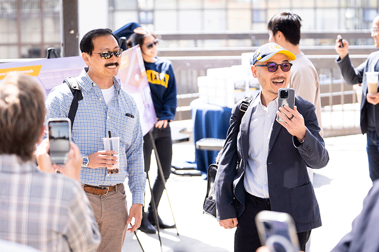 Ken is wearing a blue and white plaid shirt and holding a boba tea. Coleman is wearing a Cal cap and dark blazer. Both men are smiling and interacting with other guests.