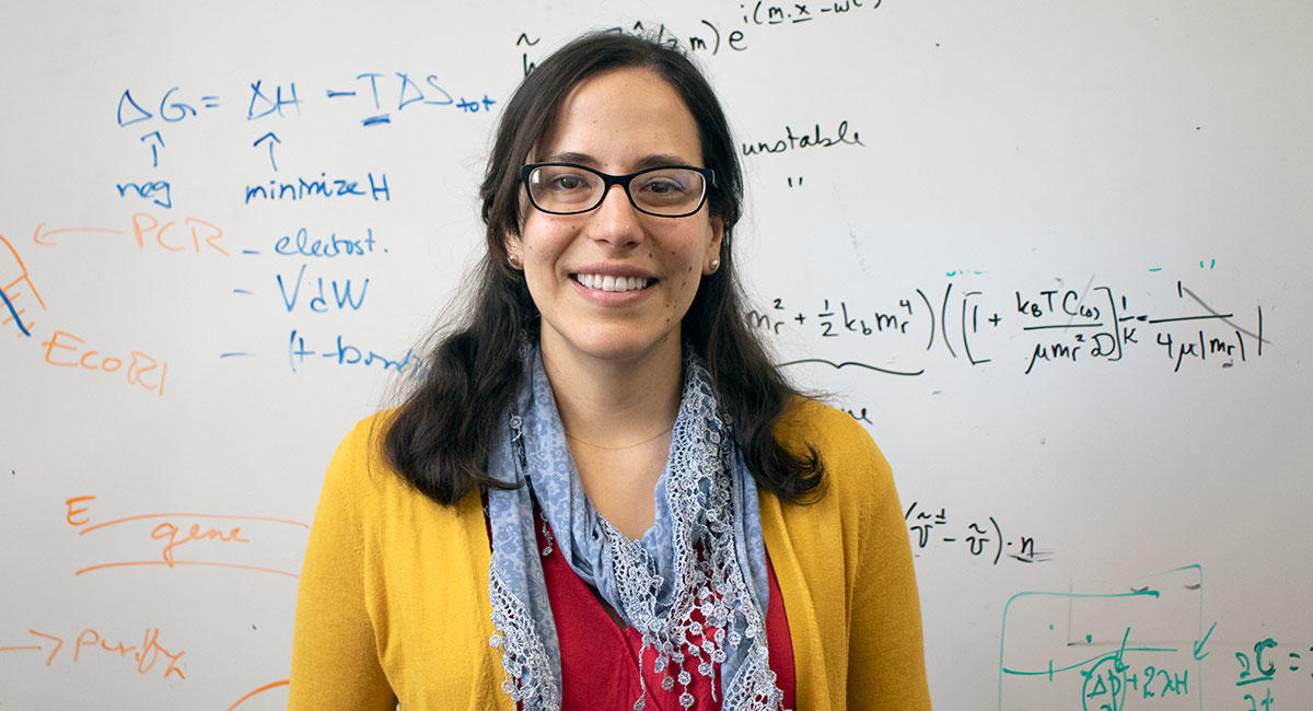 Photo of Markita in a yellow sweater, red top, and lacy blue scarf, standing in front of a whiteboard filled with equations.