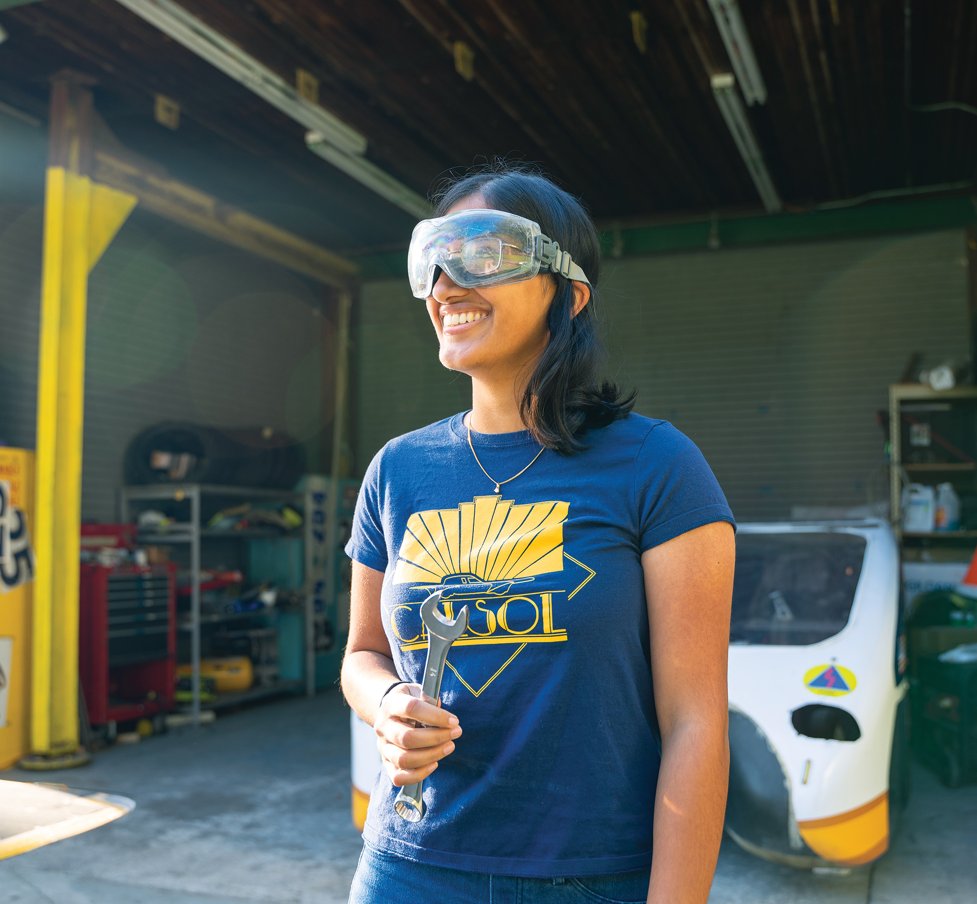 Photo of Lekha wearing a blue and gold Cal Sol tshirt, holding a wrench, and wearing goggles.