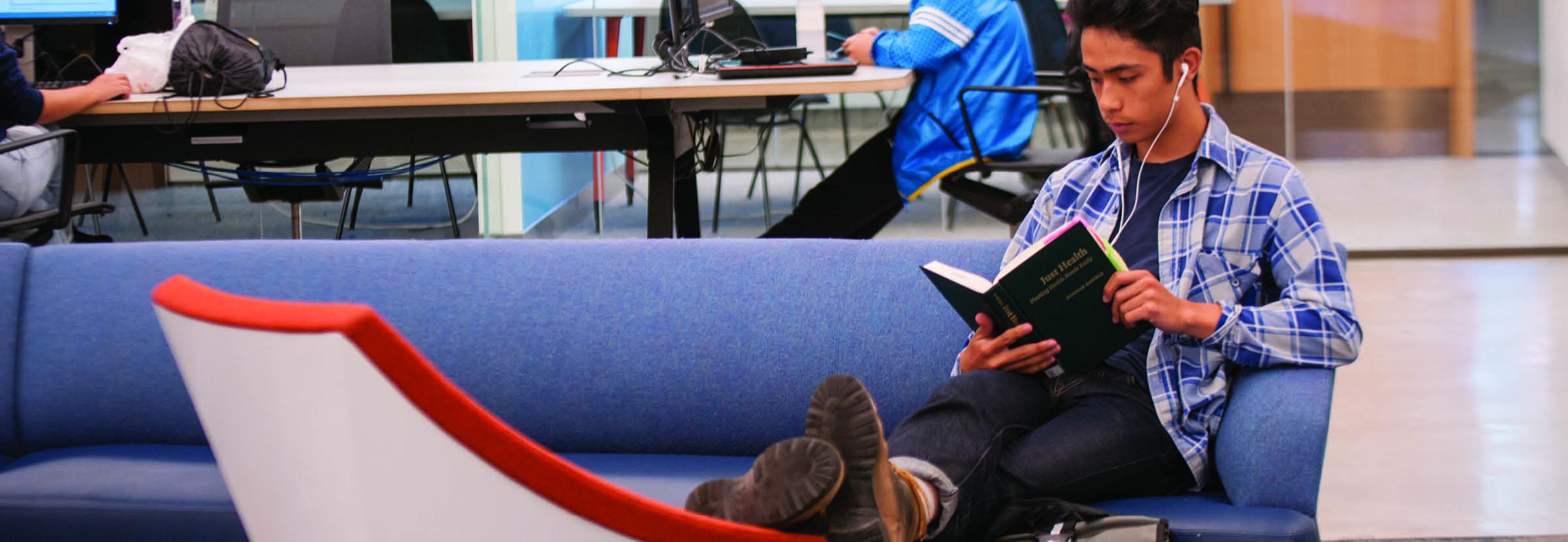 Photo of a male student in a blue plaid shirt sitting on a couch reading a textbook.