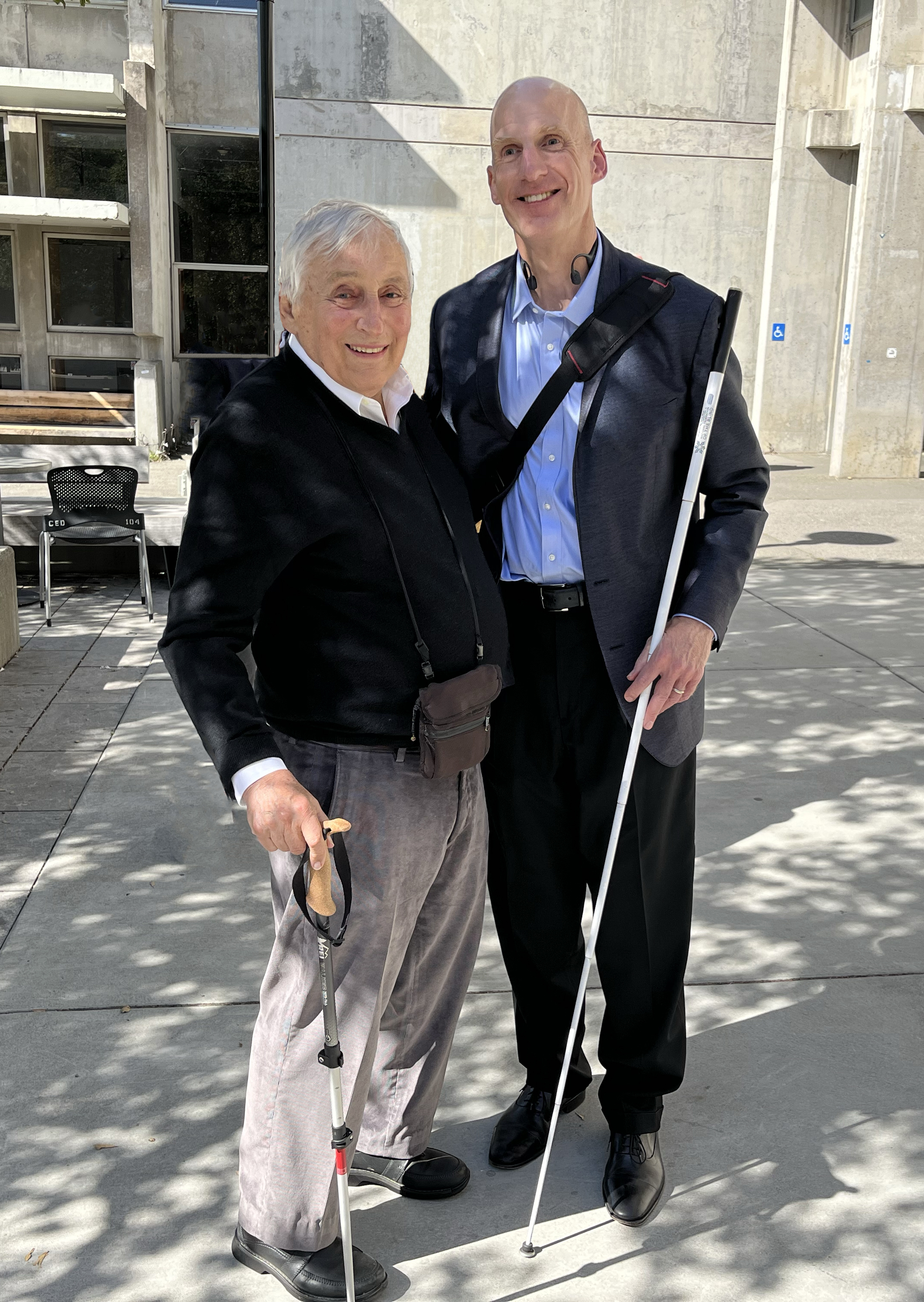 Photo of Ray, wearing gray pants and a dark sweater, standing with Chris, who is blind and uses a cane.