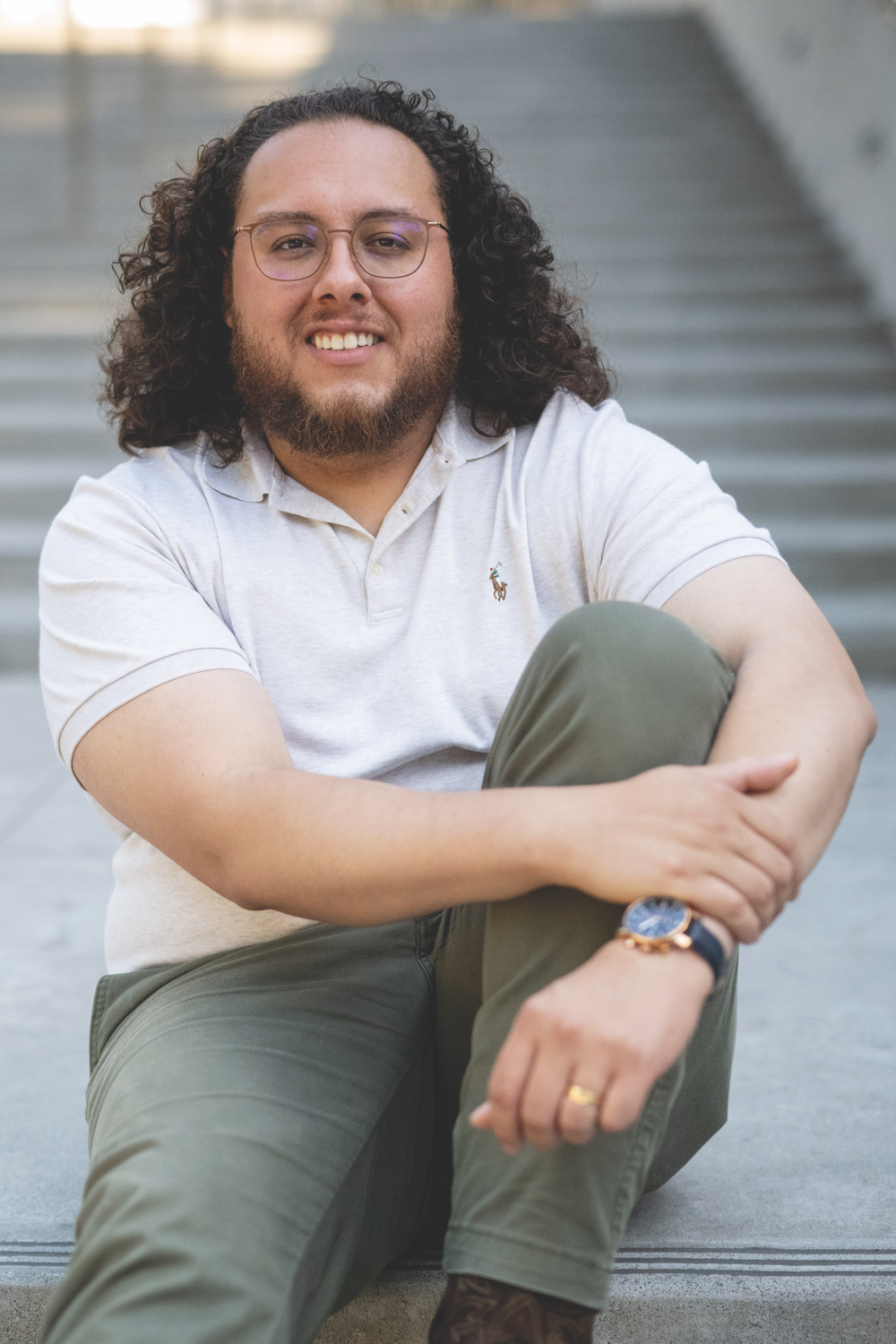 Photo of Luis in a white polo shirt and olive green pants, sitting on concrete stairs.