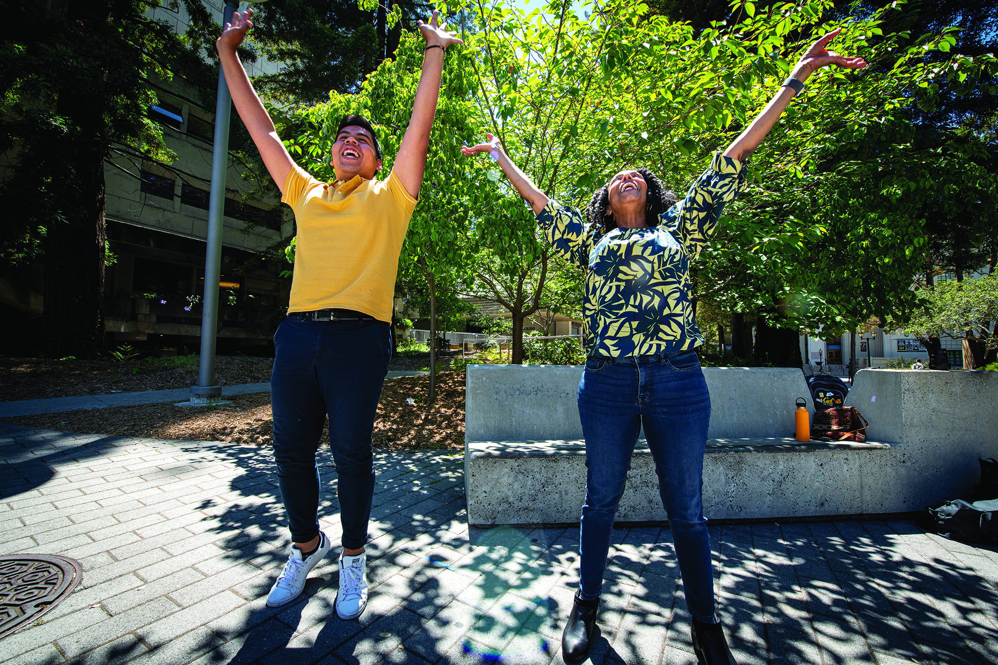 Photo of Joseph in a bright yellow shirt and Margo in blue and yellow floral shirt exuberantly jumping with their arms outstretched.