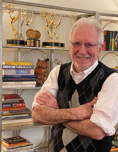 Color photo of Mark wearing a white shirt and black and gray argyle sweater, standing in front of a bookshelf lined with trophies.