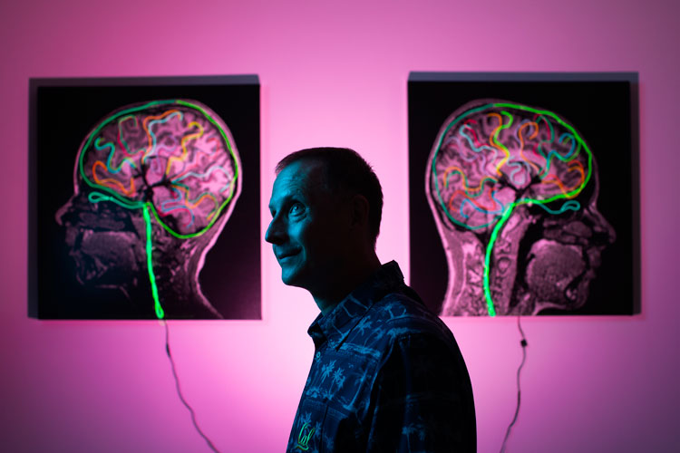 Photo of Michael Silver, partially lit by blue light and partially in shadow, with two digital representations of brains lit up in pink on a pink wall behind him.