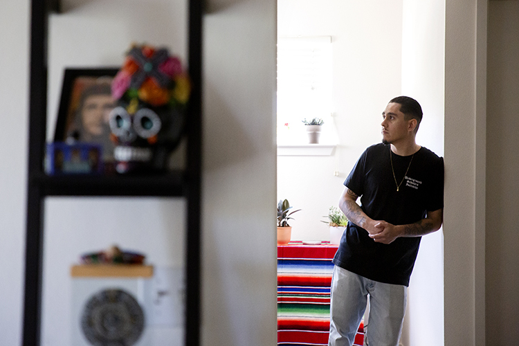 Photo of a young man standing in the hallway of his home, Mexican blanket and a small plant on a table behind him, decorative calavera statue and framed photo of Che Guevara blurry in the foreground