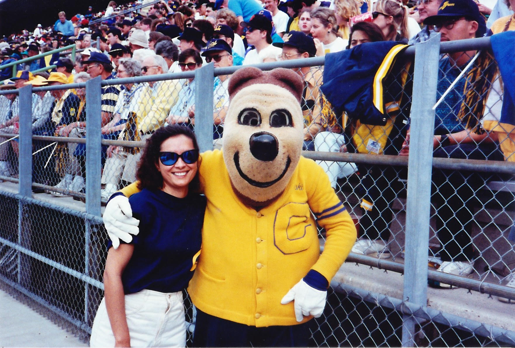 Photo of Michele and Oski in front of the stands. He's wearing his famous letter sweater and white gloves. She's wearing white shorts and a dark, sleeveless top.