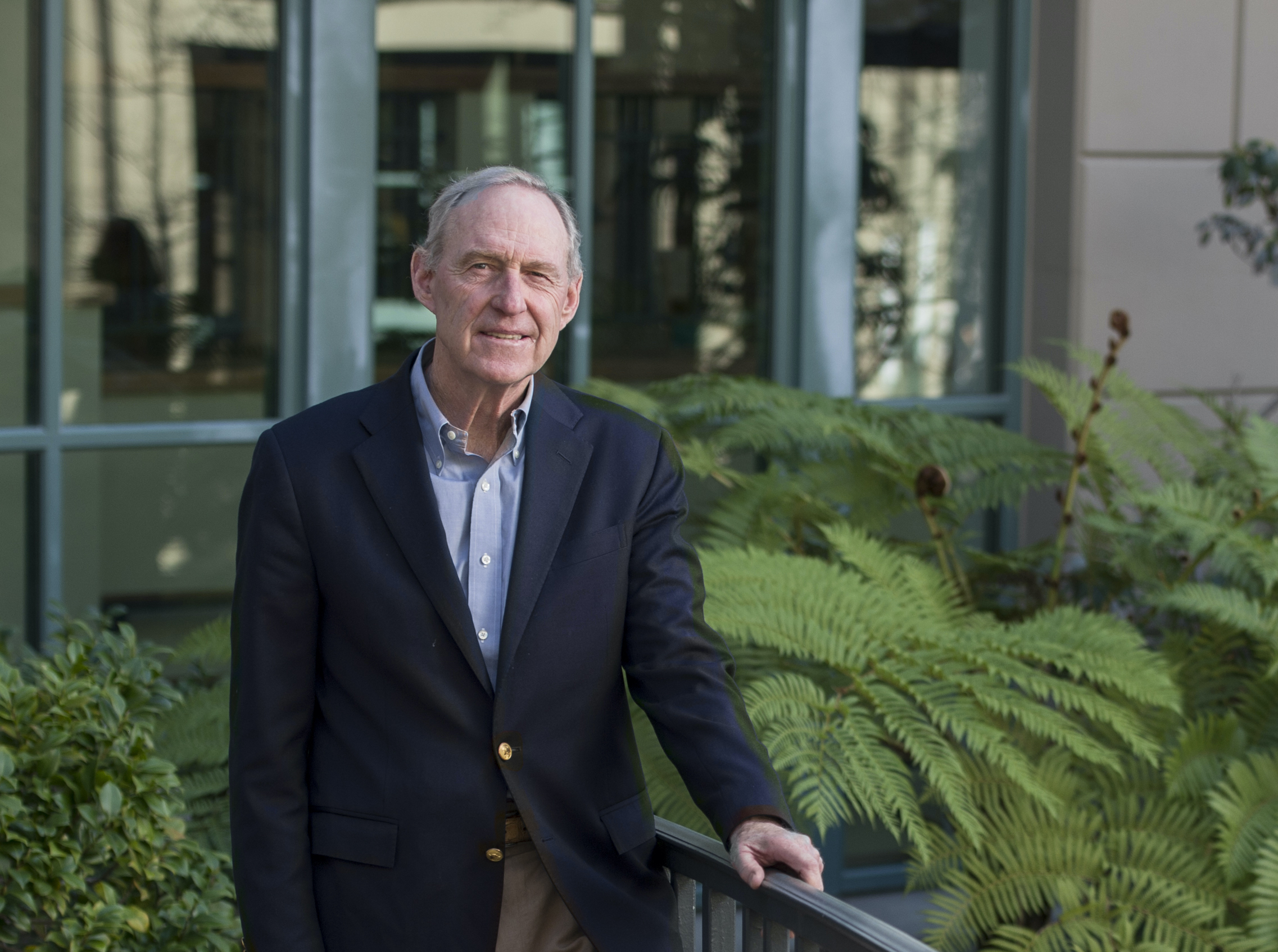 Photo of Ned in khaki pants, a light blue shirt, and dark blue blazer standing against a railing with large ferns and other shrubbery in the background.