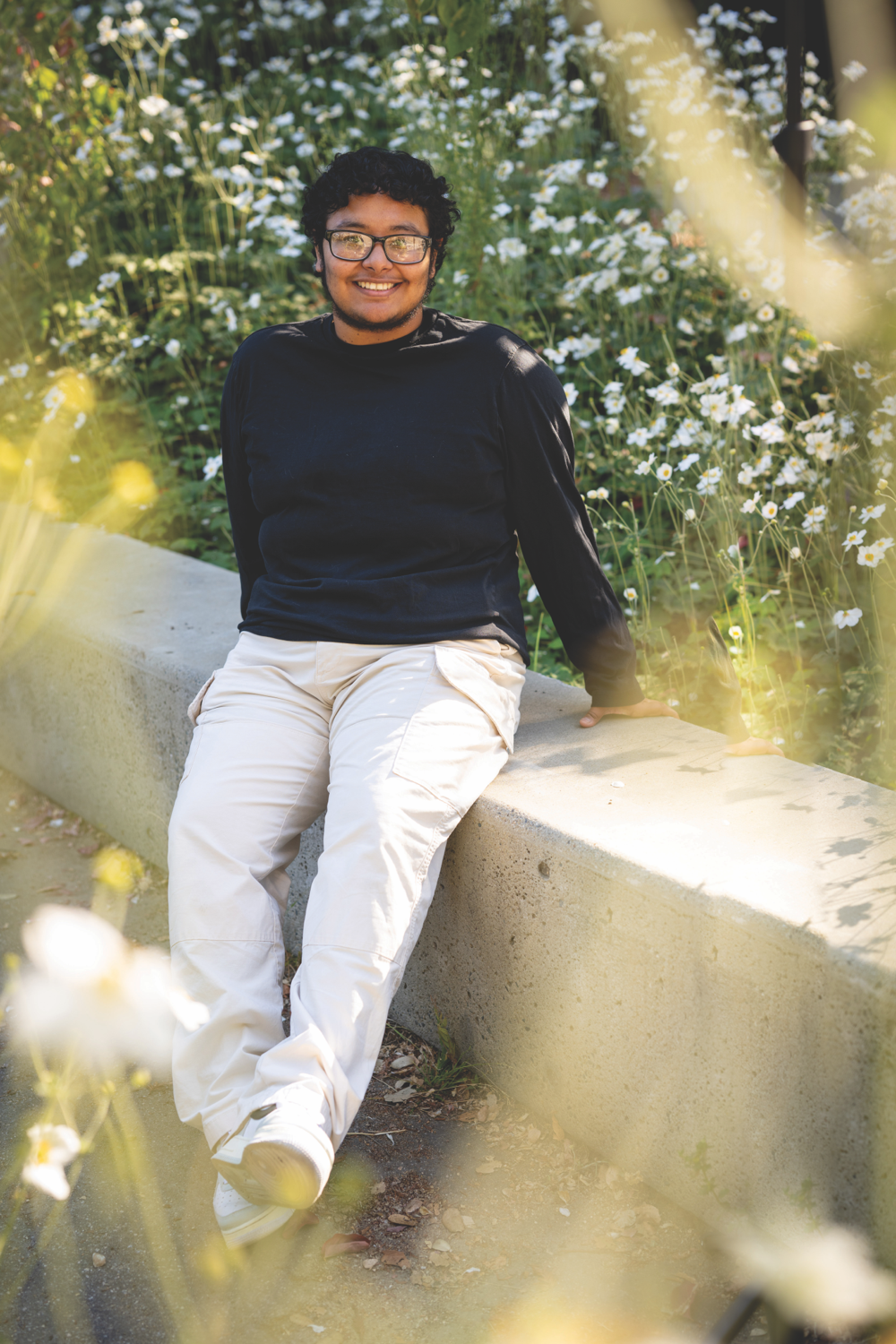 Photo of Nilo in light-colored pants and long-sleeved black top, sitting on a concrete wall by white flowers.