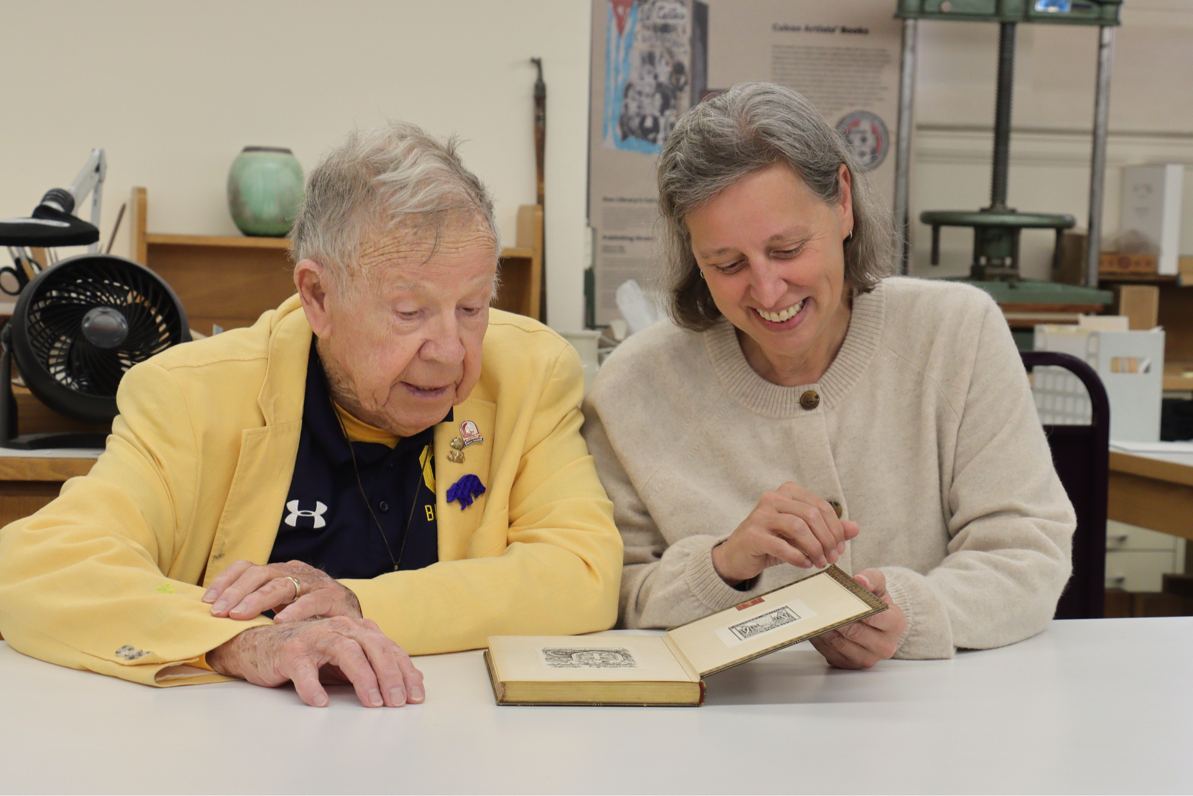 Photo of Peter wearing a yellow blazer and Erika in a cream sweater bent over an older book with illustrations.