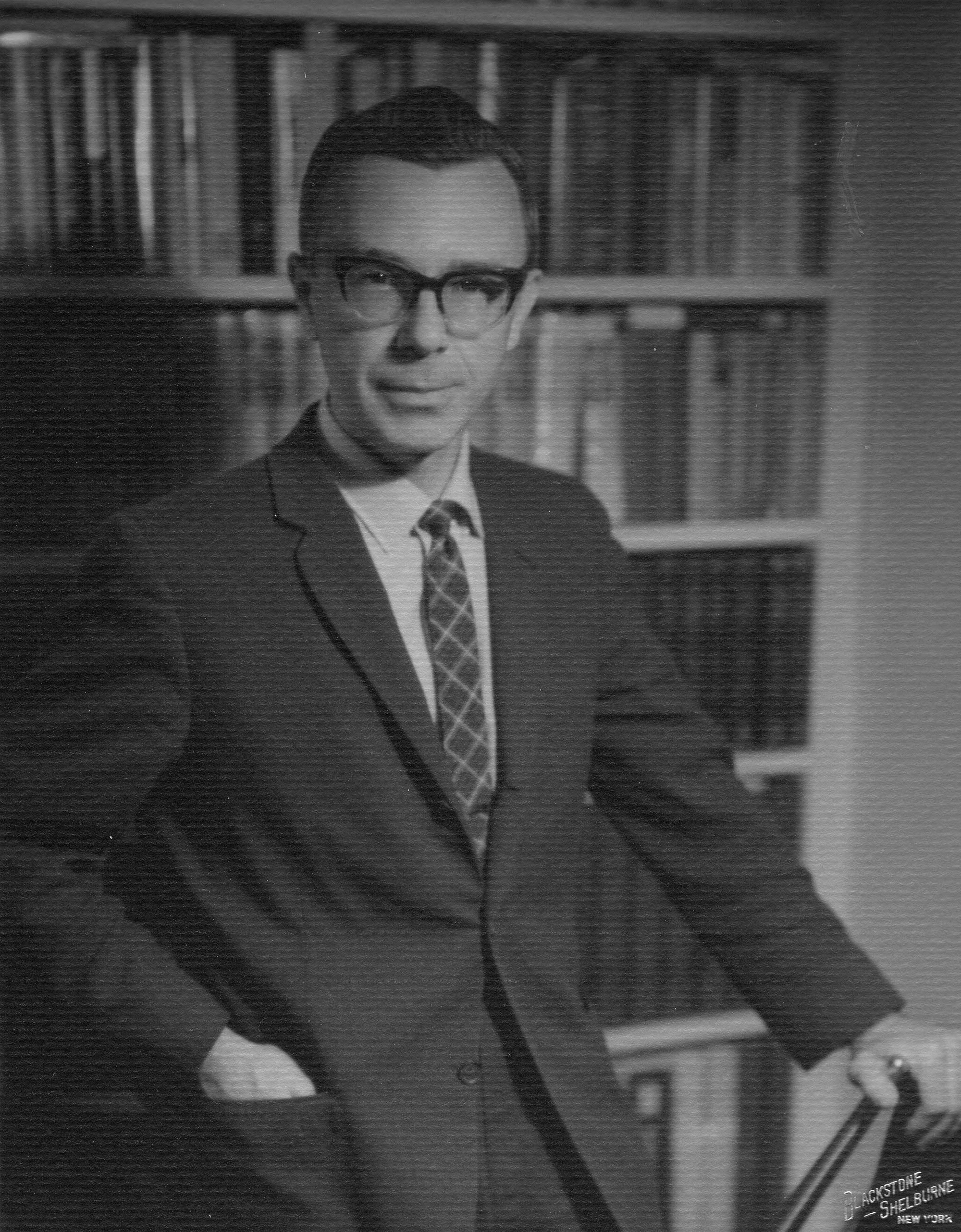 Photo of Robert circa mid-1960s in a coat and tie standing by a bookshelf