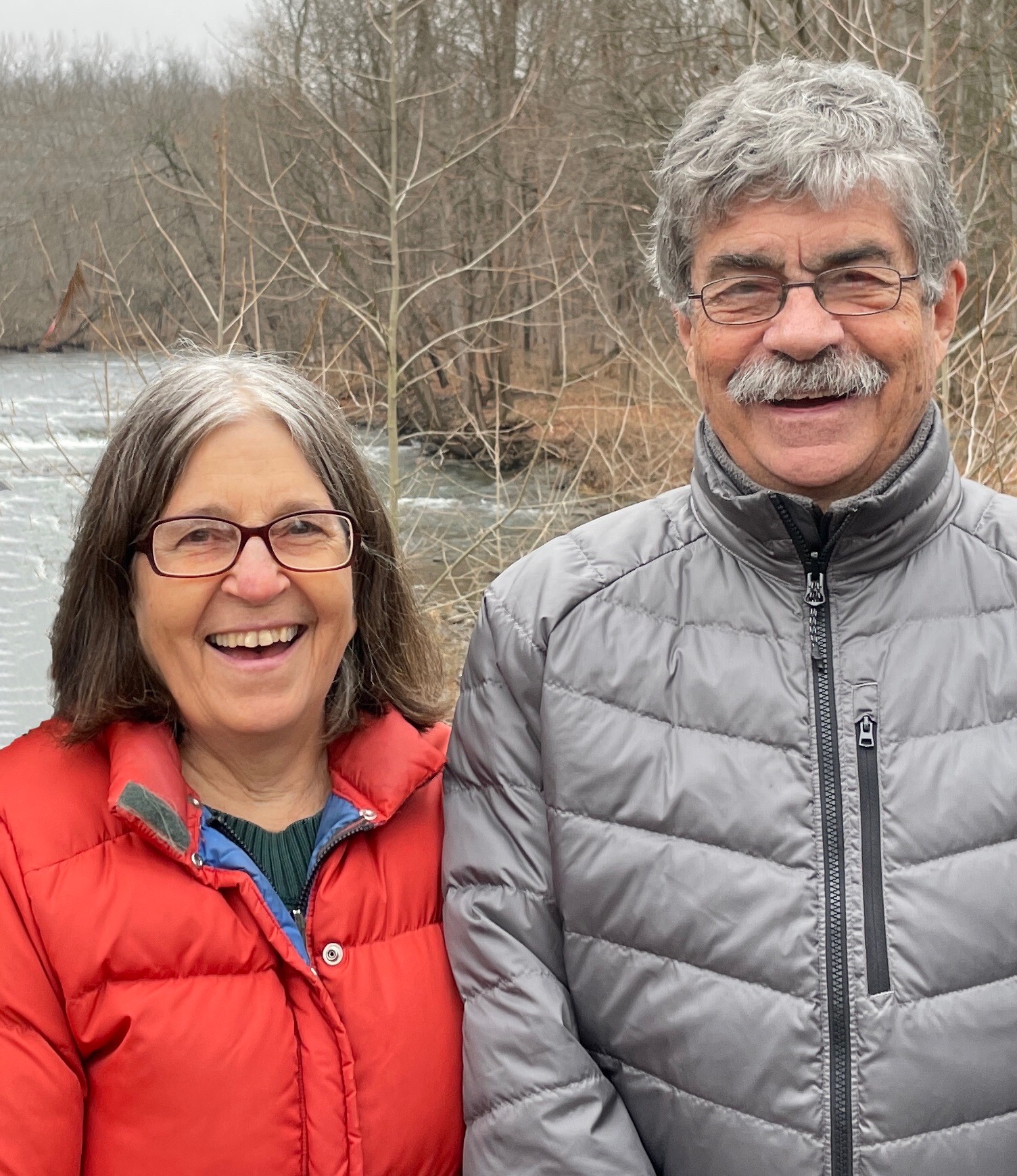 Color photo of two people outside, wearing puffy jackets, smiling at the camera