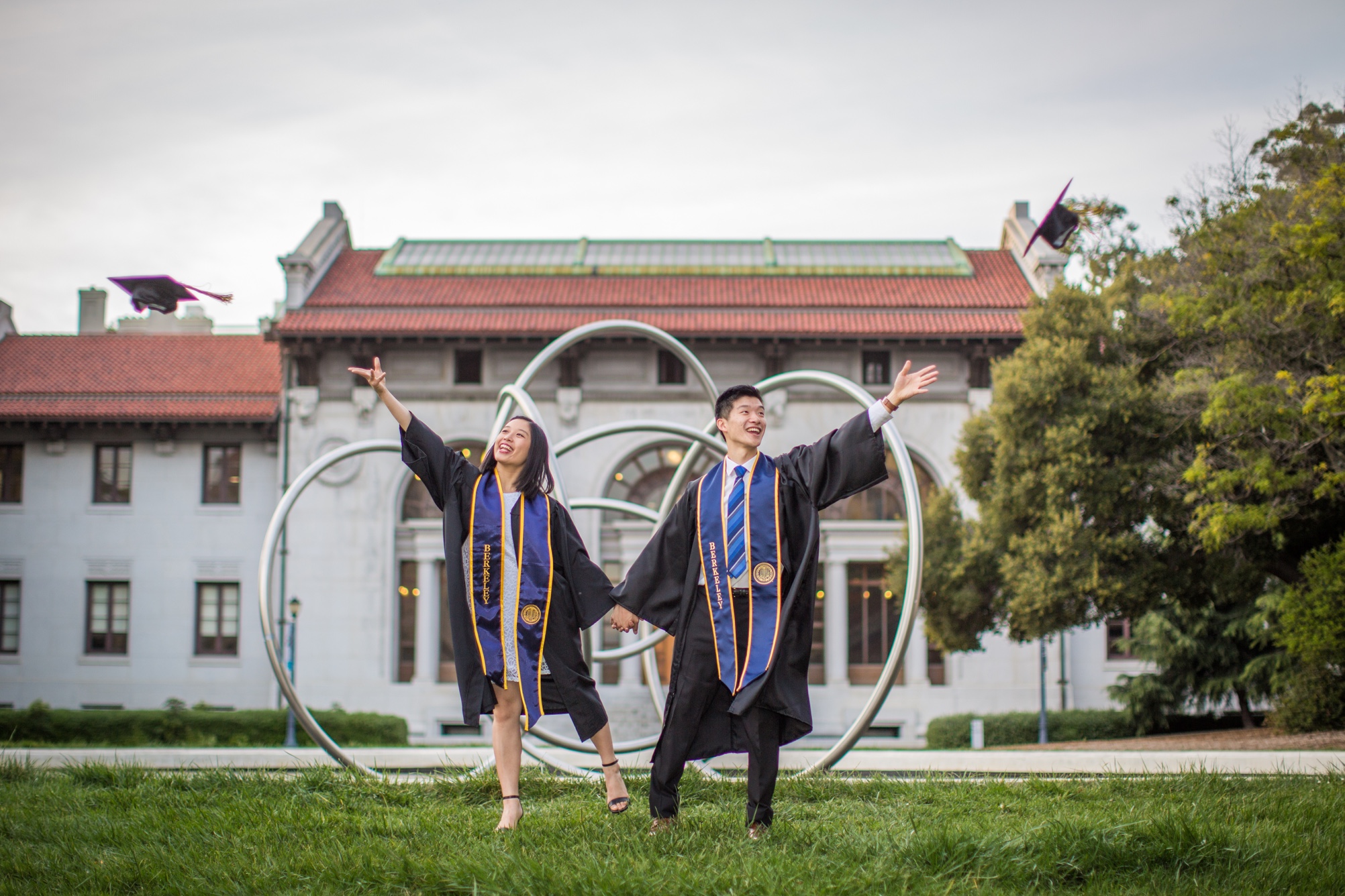Photo of Crystal and Robert in graduation regalia throwing their caps in the air in front of 
