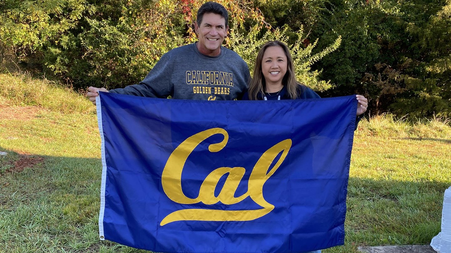 Photo of Ron and Stephanie Rivera, holding a bright blue-and-gold Cal flag in front of them, standing on a green field.