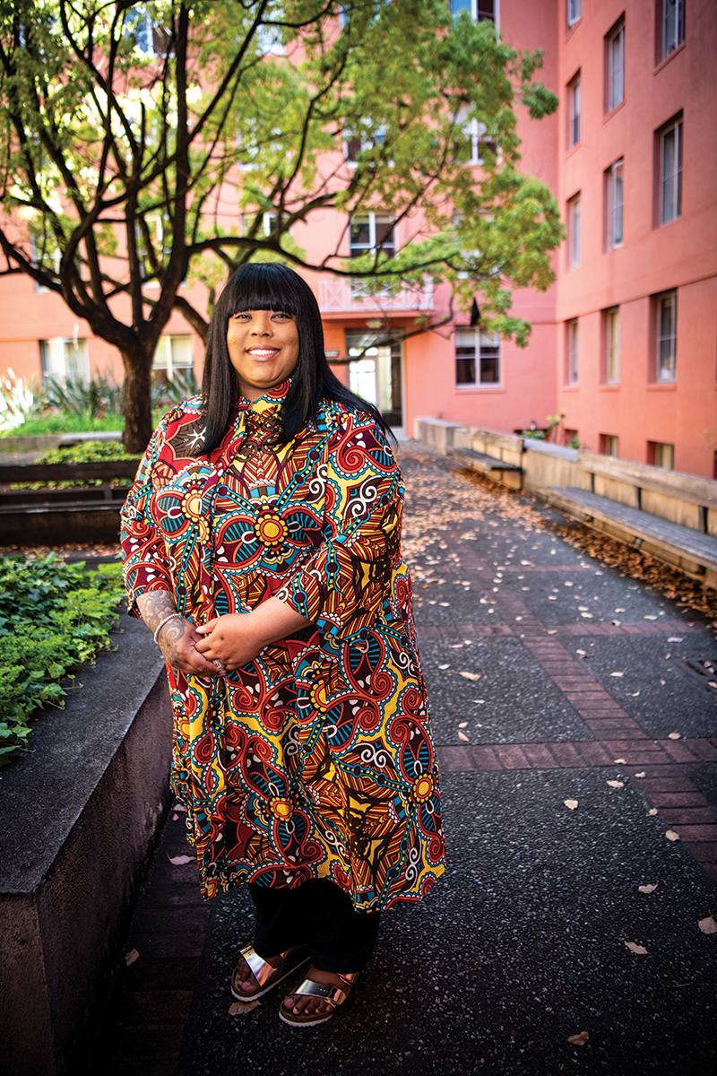 Photo of Stephanie, a Black woman, standing in a courtyard and wearing a red, yellow, and turquoise patterned dress.